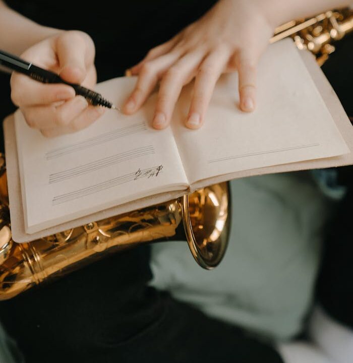 Person writing music in a notebook while holding a saxophone, capturing creativity.
