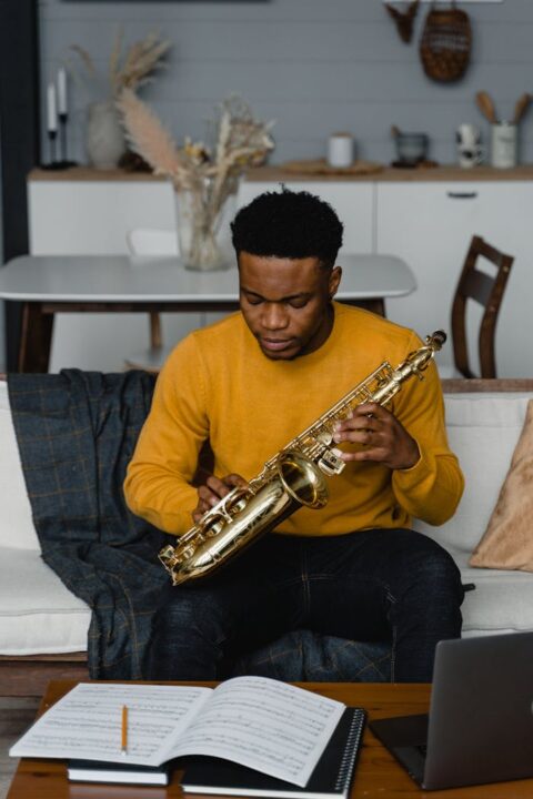 A young man sits on a couch playing a saxophone, surrounded by sheet music.